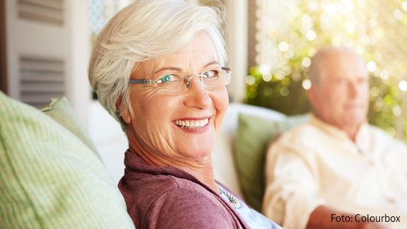Foto einer alten Frau, die auf einem Sofa sitzt und lächelnd in die Kamera blickt.