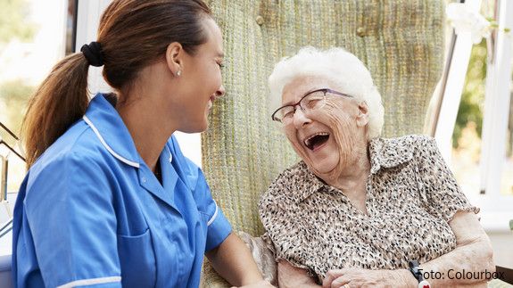 Szene aus Seniorenresidenz: Eine Seniorin sitzt auf einer Bank, neben ihr eine Pflegefachkraft. Beide lachen.
